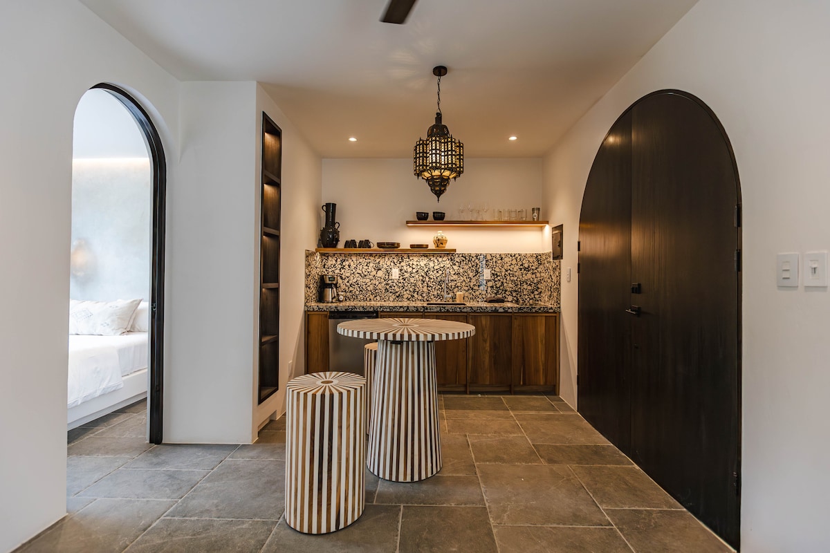 A modern kitchenette area features intricate tile work and wooden cabinetry, illuminated by soft lighting. A round table with two striped stools invites casual dining. Curved archways create an open and inviting atmosphere, with a glimpse of the bedroom area in the background.