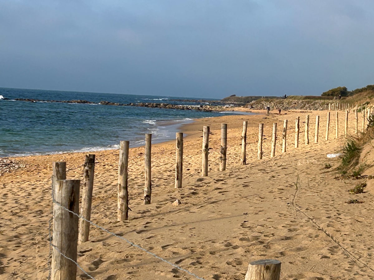 A sandy beach stretches along the coastline, lined with vertical wooden posts that mark the boundary. The calm waves gently lap against the shore, while the distant horizon meets a partly cloudy sky. The natural landscape offers a serene atmosphere for visitors.