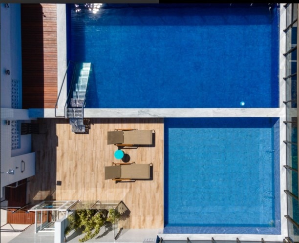 An aerial view captures two swimming pools side by side, surrounded by wooden decking. Sun loungers and a small circular table are positioned nearby, with greenery visible in the corner, creating a serene outdoor space for relaxation.