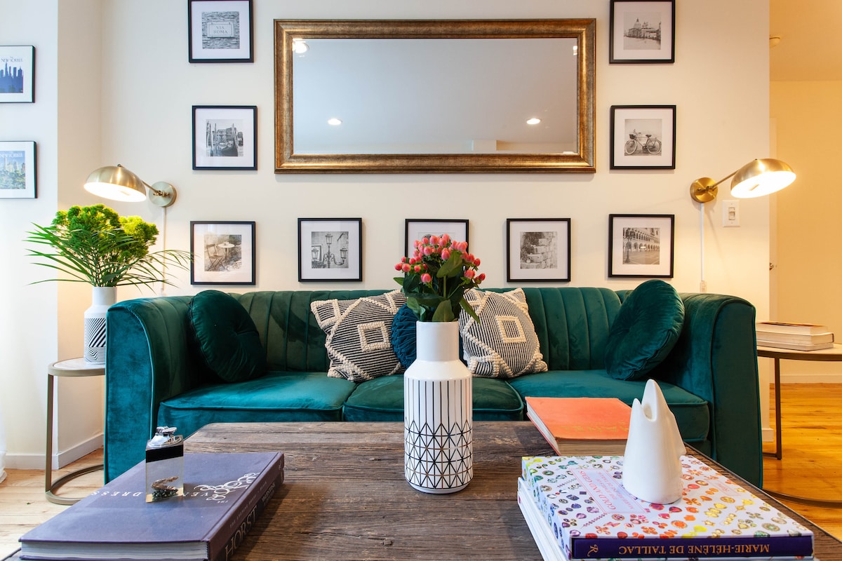A stylish living area features a vibrant green velvet sofa adorned with decorative pillows, set against a gallery wall of framed black-and-white photographs. A central wooden coffee table displays various books and a white vase filled with flowers, complemented by warm lighting from modern sconces.