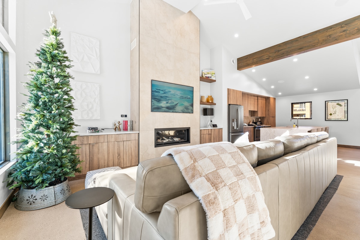 A spacious living area features a modern sofa draped with a cozy throw, positioned near a stone fireplace and a wall-mounted flat-screen TV. A holiday tree stands nearby. The open layout connects to a well-equipped kitchen in the background.