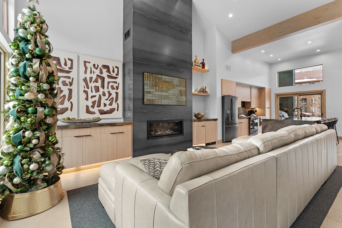 A contemporary living area features a cream-colored sectional sofa positioned near a sleek gas fireplace. A decorated Christmas tree stands in the corner, complemented by abstract wall art. Modern cabinetry is visible in the background, with stainless steel appliances in the kitchen area.