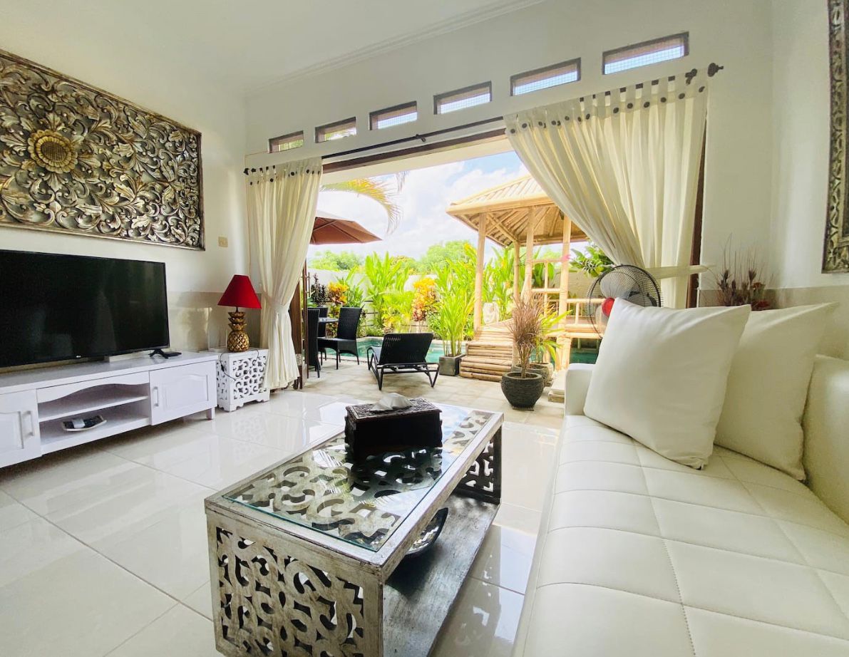 A bright living area is depicted, featuring a comfortable white sofa opposite a modern TV stand. Large glass doors open to an outdoor space, framed by elegant curtains. A decorative coffee table adds a touch of elegance, blending with the serene greenery visible through the doors.