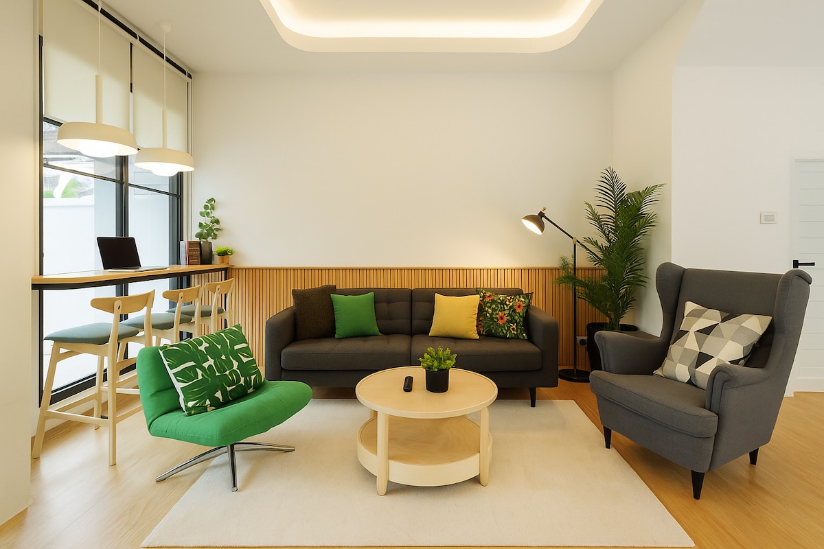 A modern living area is designed with light wood accents and a neutral color palette. Comfortable seating includes a sofa with colorful cushions and an accent chair. A circular coffee table is positioned in the center, while a dining space is visible in the background.