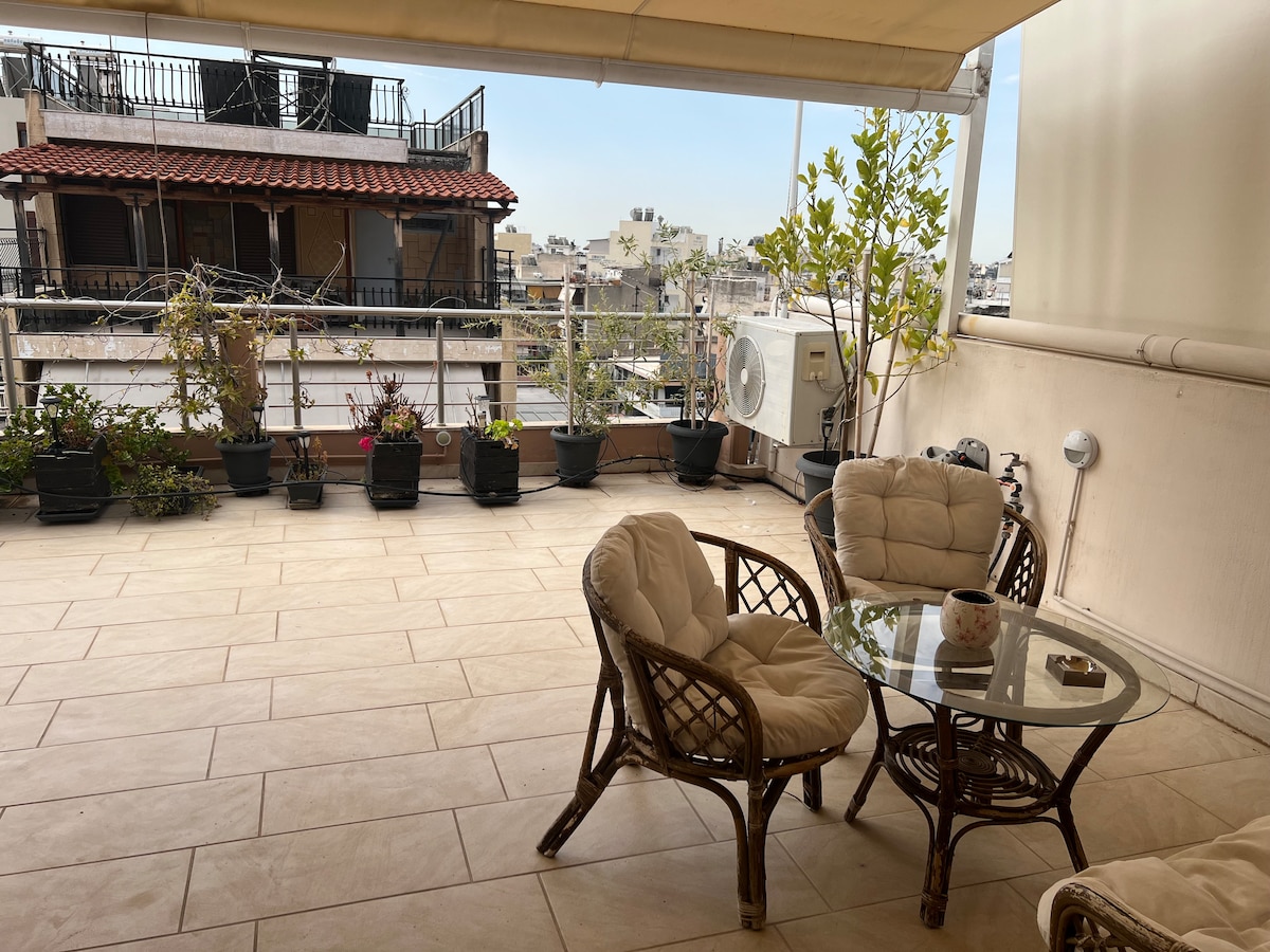 A spacious terrace features a glass-top table surrounded by two wicker chairs. Potted plants line the balcony, adding greenery to the space. A nearby air conditioning unit is visible, with a view of neighboring structures and skyline in the background.