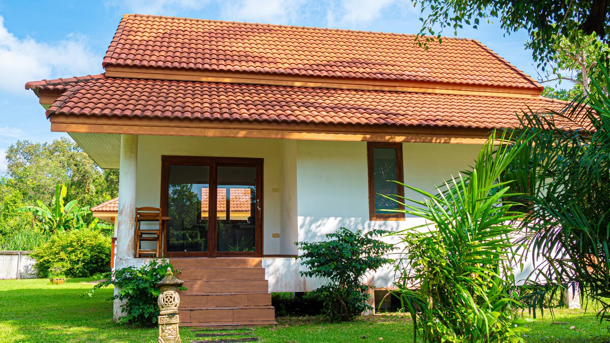 A villa exterior is featured, showcasing a red-tiled roof and large windows, surrounded by lush greenery. The entrance includes a small staircase leading to a spacious porch, inviting guests to relax in the natural setting.