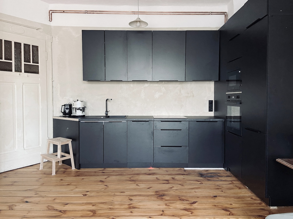 A modern kitchen features dark cabinets with sleek finishes and a light wood floor. Essential appliances, including a coffee maker and oven, are visible. A small wooden step stool stands to the side, providing additional convenience in the space.
