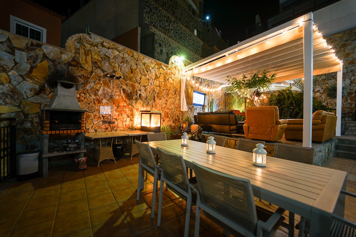 An outdoor dining area is set under a pergola, featuring a long table surrounded by chairs. Soft lighting illuminates the space, creating a cozy atmosphere. A stone wall and grill are visible in the background, enhancing the outdoor experience.