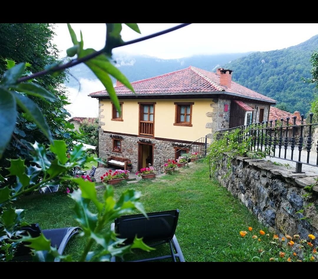 A rustic two-story building is surrounded by greenery, featuring a textured stone facade and vibrant yellow walls. The sloped roof is topped with red tiles. A well-kept garden area includes flower beds and overlooks a mountainous landscape, offering a peaceful outdoor retreat.