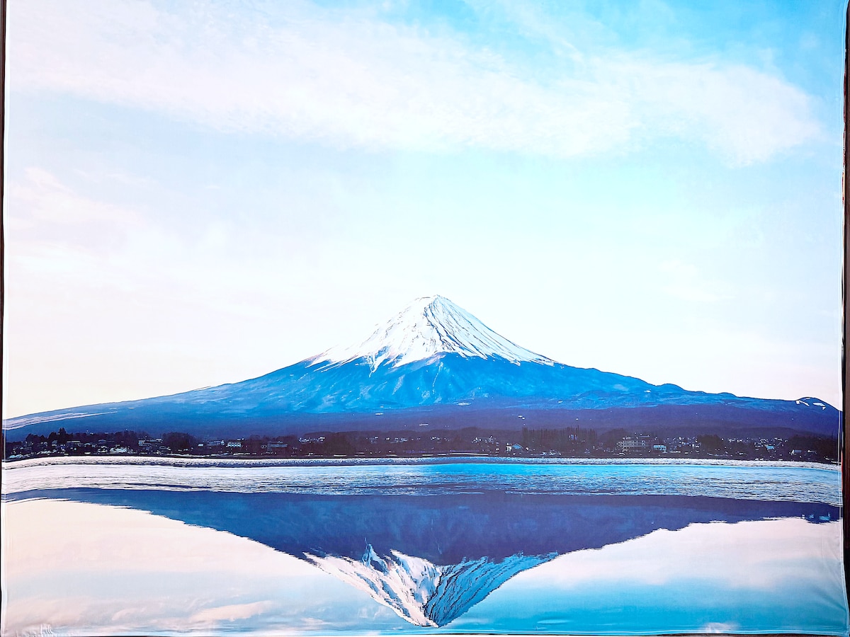 A serene landscape captures the majestic Mt. Fuji, its snow-capped peak reflected in calm waters below. The sky is painted in soft shades of blue and white, enhancing the tranquil scene that invites admiration for the natural beauty surrounding the area.