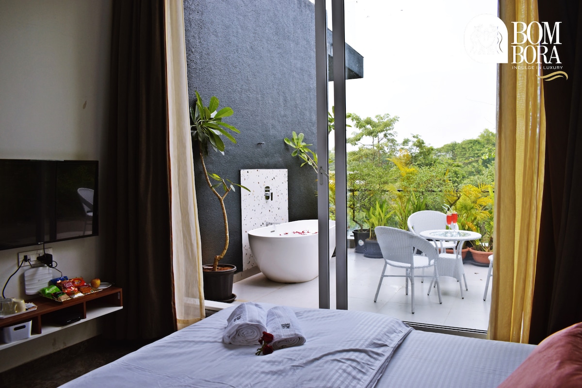 A bright and airy bedroom features a spacious bed with neatly arranged towels. Floor-to-ceiling windows offer a view of an outdoor patio with a bathtub and seating area surrounded by lush greenery.