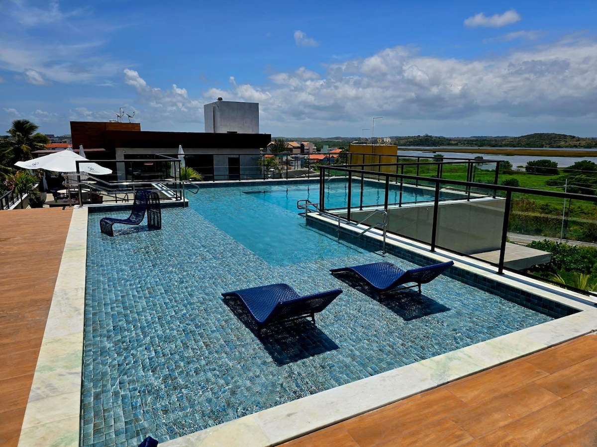 A spacious rooftop area features a large swimming pool with clear blue water. Two lounge chairs are positioned beside the pool, while the surrounding deck is lined with modern landscaping. A view of lush greenery and distant buildings can be seen in the background under a bright sky.
