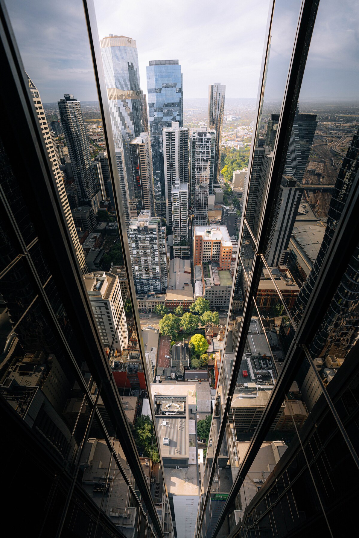 A breathtaking view from the 61st floor showcases a vibrant city landscape. Towering skyscrapers reflect light and feature a mix of architectural styles. Below, a tree-lined street is visible, providing a contrast to the urban environment, with the horizon extending into the distance.