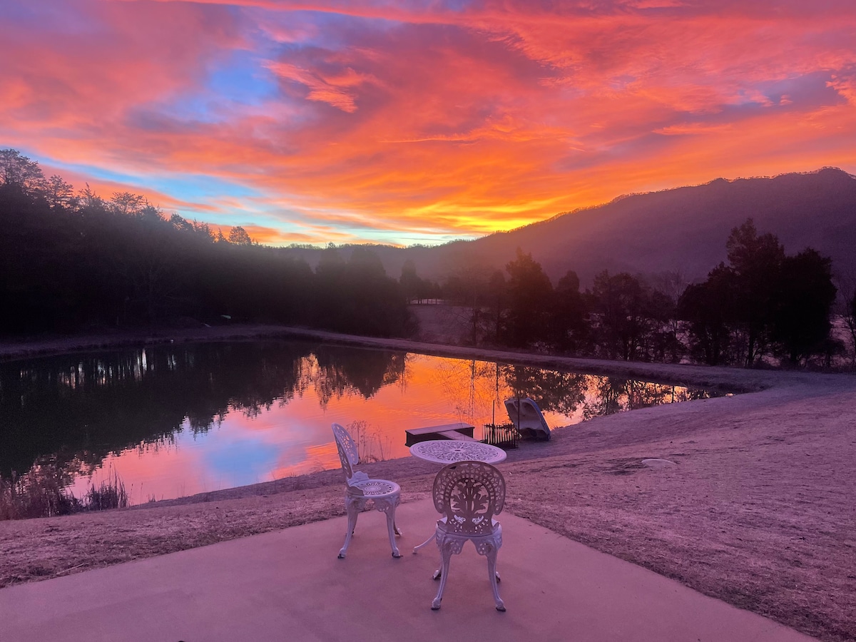 Two white chairs are positioned beside a tranquil pond, reflecting a vivid sunset with shades of pink, orange, and purple. The surrounding landscape features trees and mountains in the background, creating a serene outdoor space for relaxation.