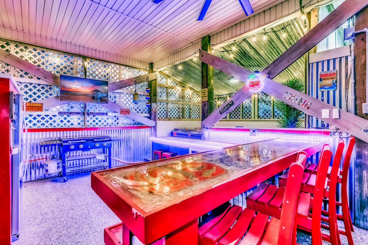An enclosed entertainment area features a large bar with red stools arranged around it. Bright lighting is reflected off the tables, creating a lively atmosphere. A grill is positioned against the wall, and a television screen is mounted for viewing. Decorative details enhance the space.