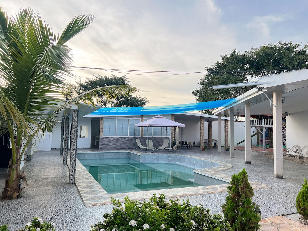 A spacious outdoor area features a tranquil pool surrounded by decorative tiles. A shaded seating area is available under a blue canopy, with lounge chairs positioned nearby. Lush green plants provide a natural border, enhancing the inviting setting for relaxation.