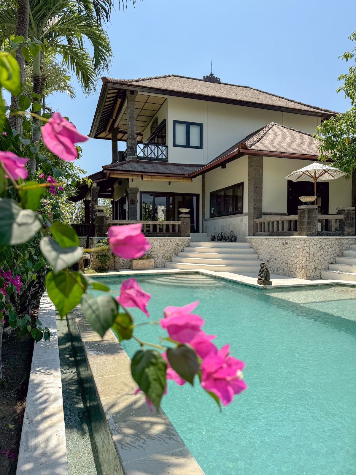 The exterior of the villa is framed by vibrant pink bougainvillea flowers. A refreshing lap pool glistens under the sun, leading to the entrance of the two-story structure, which features traditional Balinese architecture and shaded outdoor seating areas.
