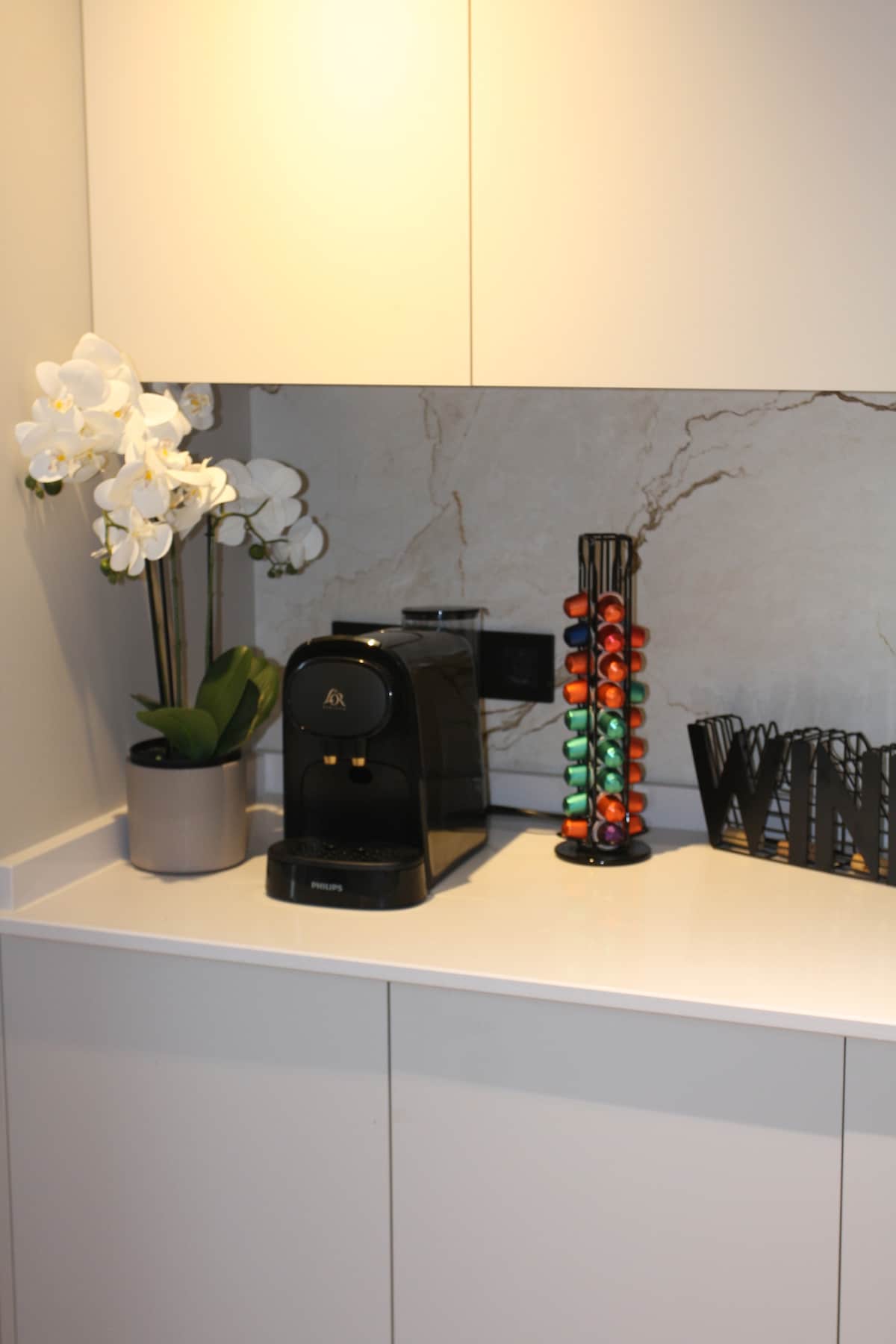 A modern kitchen area includes a sleek coffee machine positioned next to a colorful capsule holder. A potted orchid adds a touch of nature, contrasting with the neutral cabinetry and marble backsplash.