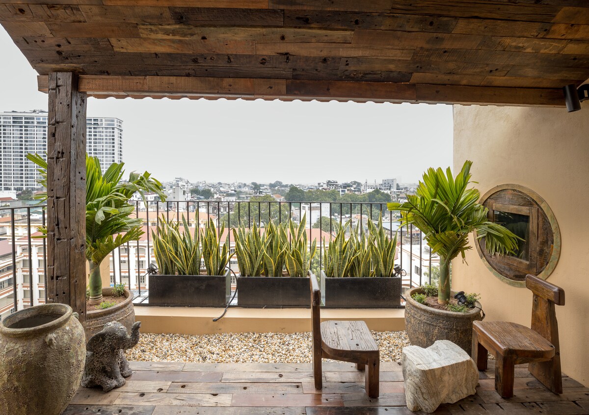A serene balcony space is highlighted by two wooden chairs and a rustic bench, surrounded by potted plants. The view features rooftops and distant buildings, framed by a protective railing. The warm wood tones and natural elements create a calming outdoor retreat.
