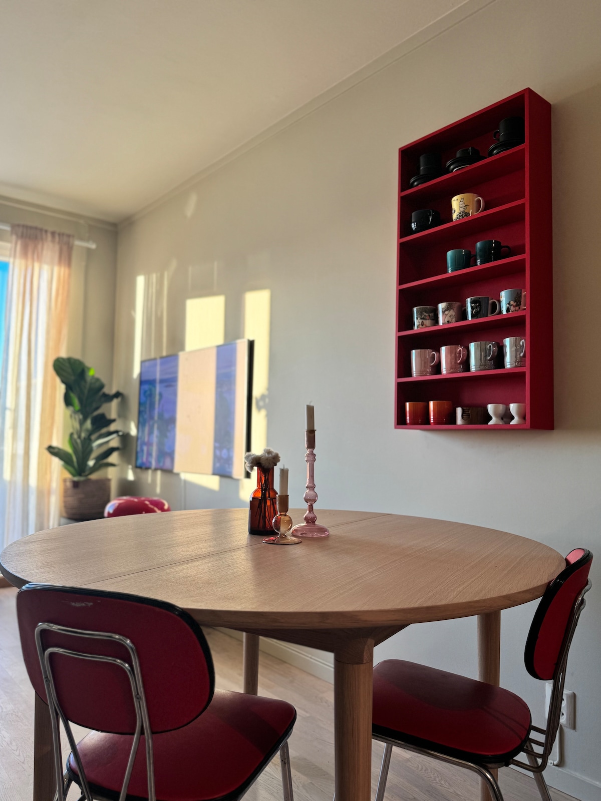 A round dining table is centered in a sunlit area, accompanied by red chairs. A shelf in vibrant red displays an assortment of colorful mugs and decorative items. A tall plant adds a touch of greenery, while a large television is mounted on the wall.