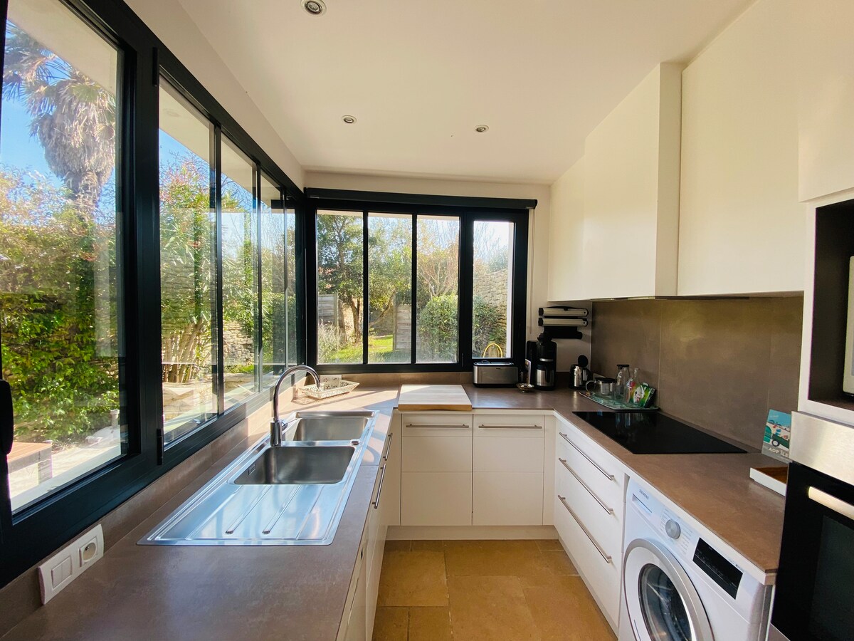 A modern kitchen features sleek white cabinetry and countertops, with large windows providing views of the garden. An induction cooktop and an oven are visible, along with essential appliances including a microwave and a washing machine. Natural light fills the space.