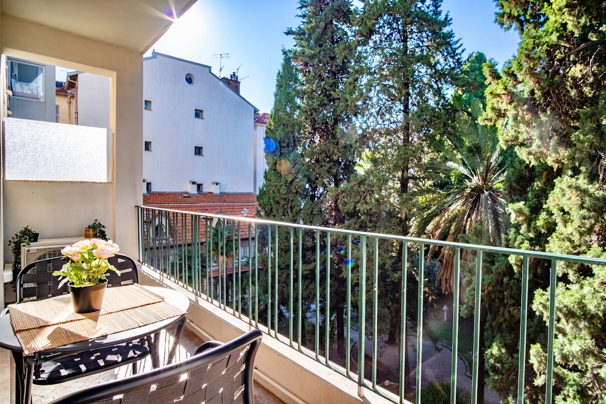 A balcony is featured with a small table and four chairs, overlooking a tranquil garden area filled with lush trees. Natural light filters through, creating a serene outdoor space that invites relaxation.