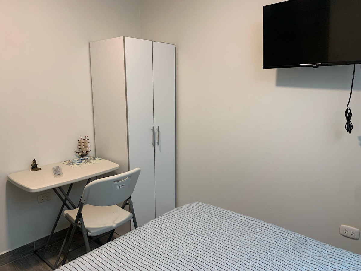 A simple room is presented with a compact bed and a wall-mounted television. A small table and chair are positioned next to a wardrobe, which provides storage space. Soft lighting enhances the minimalistic design of the space.