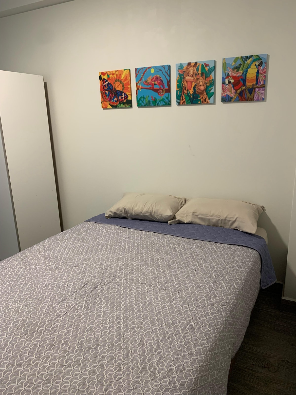A bedroom is shown with a spacious bed, covered by a light-colored quilt. Two soft pillows rest at the head of the bed. Four colorful artworks are displayed above the bed, adding a vibrant touch to the otherwise neutral room. A white wardrobe is partially visible to the left.