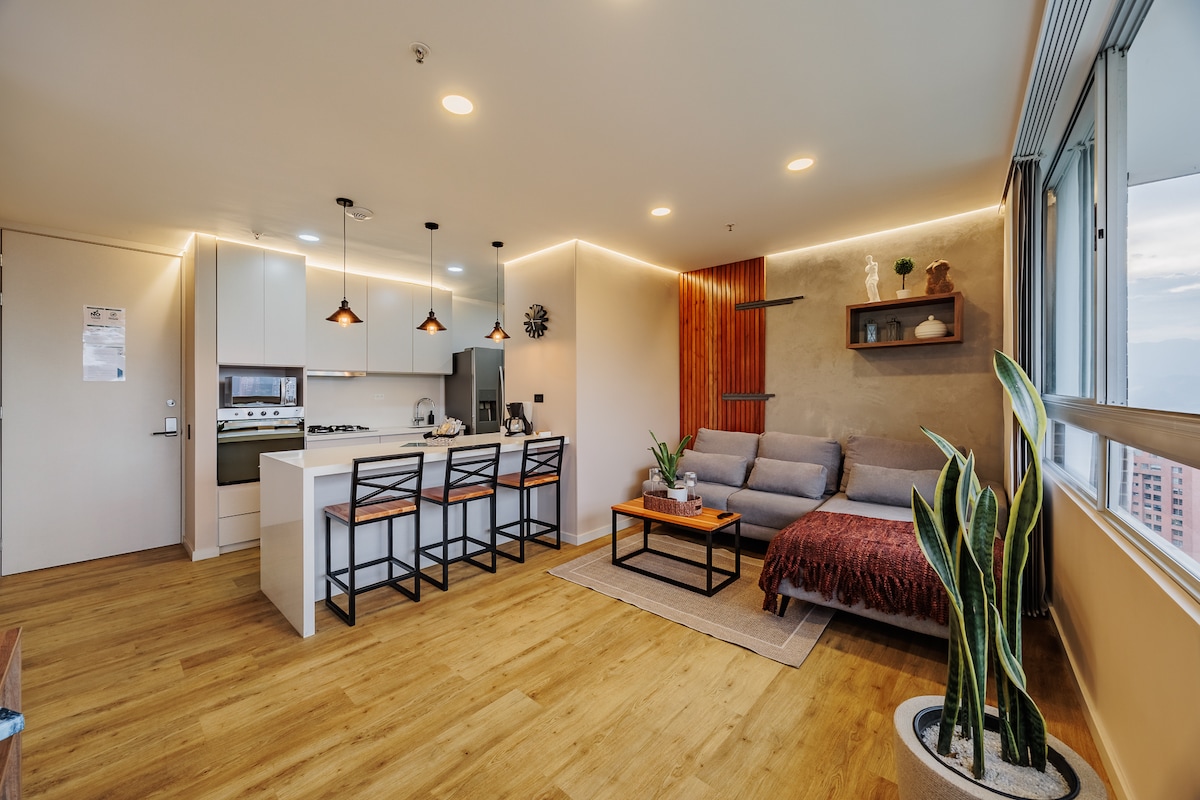A modern living space features a gray sofa and a wooden coffee table, complemented by potted plants. An open kitchen area includes white cabinetry and bar seating with four black stools. Natural light fills the room, highlighting the warm wood flooring and inviting ambiance.