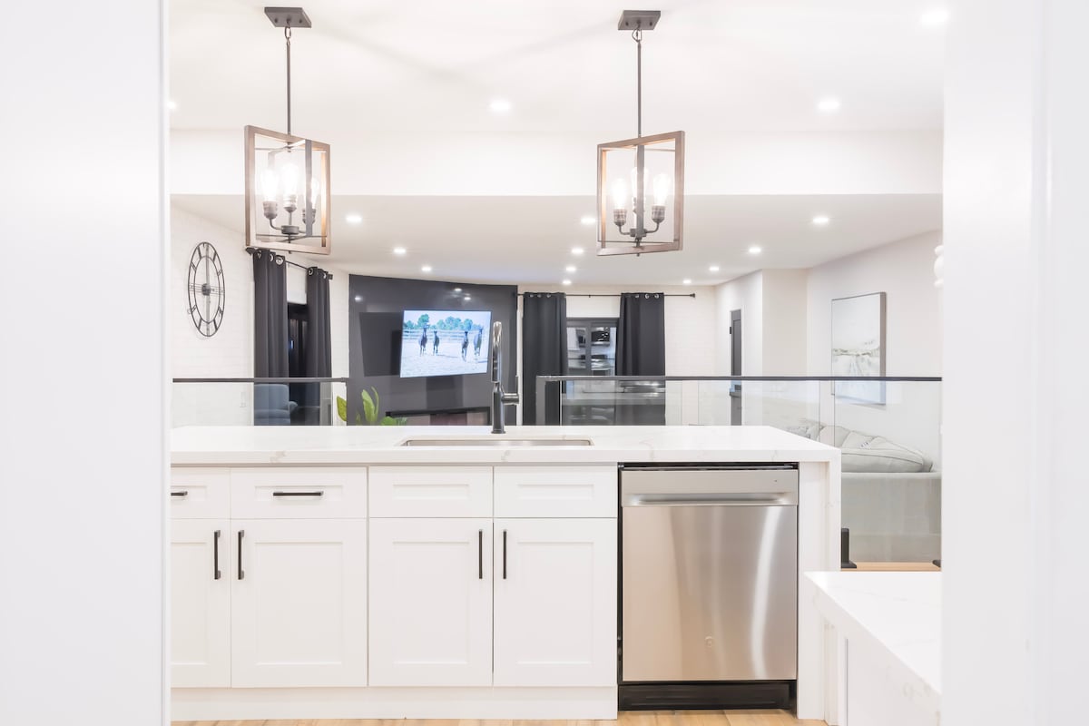 The modern kitchen features white cabinetry, a stainless steel dishwasher, and a large sink. Two contemporary light fixtures hang above the countertop, providing bright illumination. In the background, an open living area is visible, showcasing a television and a relaxed seating space with light-colored décor.
