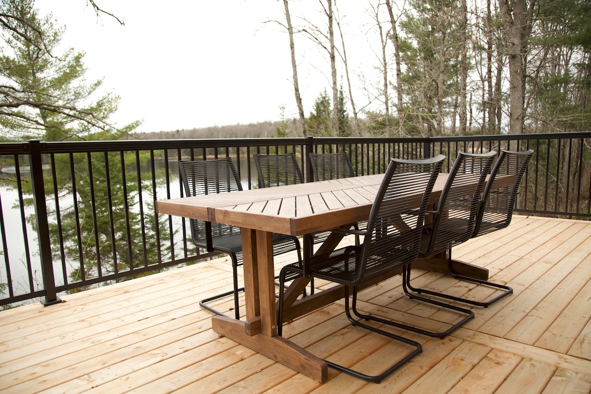 An outdoor dining area features a large wooden table with several metal chairs positioned around it, offering a view of the surrounding trees and lake. The deck is made of light-colored wood, providing a spacious setting for meals or gatherings.