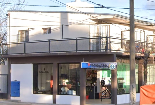 A two-story building features a modern facade with large windows and a balcony above. The entrance is framed by glass, showcasing a vibrant retail space inside. Outside, the area includes greenery and a mailbox by the sidewalk, providing a welcoming street view.
