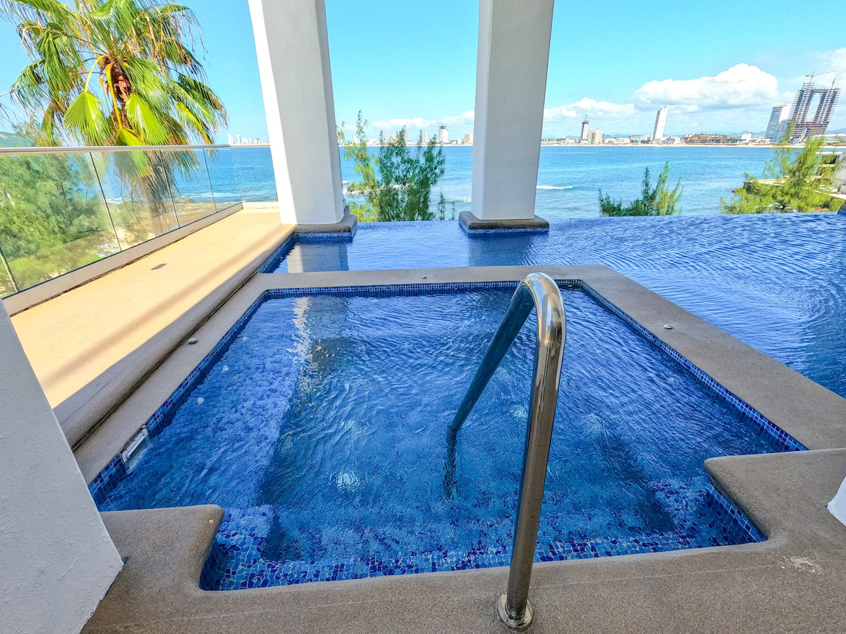 An inviting infinity pool offers a clear view of the ocean, surrounded by smooth concrete edges. The pool features steps leading into the water and is framed by lush greenery and a sunny, expansive outdoor area, creating a serene relaxation spot.