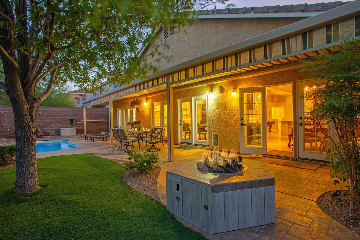 The outdoor area features a shaded patio with a dining set and grill, surrounded by lush grass. A cozy fire pit is positioned nearby, and the inviting pool glistens in the evening light, providing a warm atmosphere for gatherings.