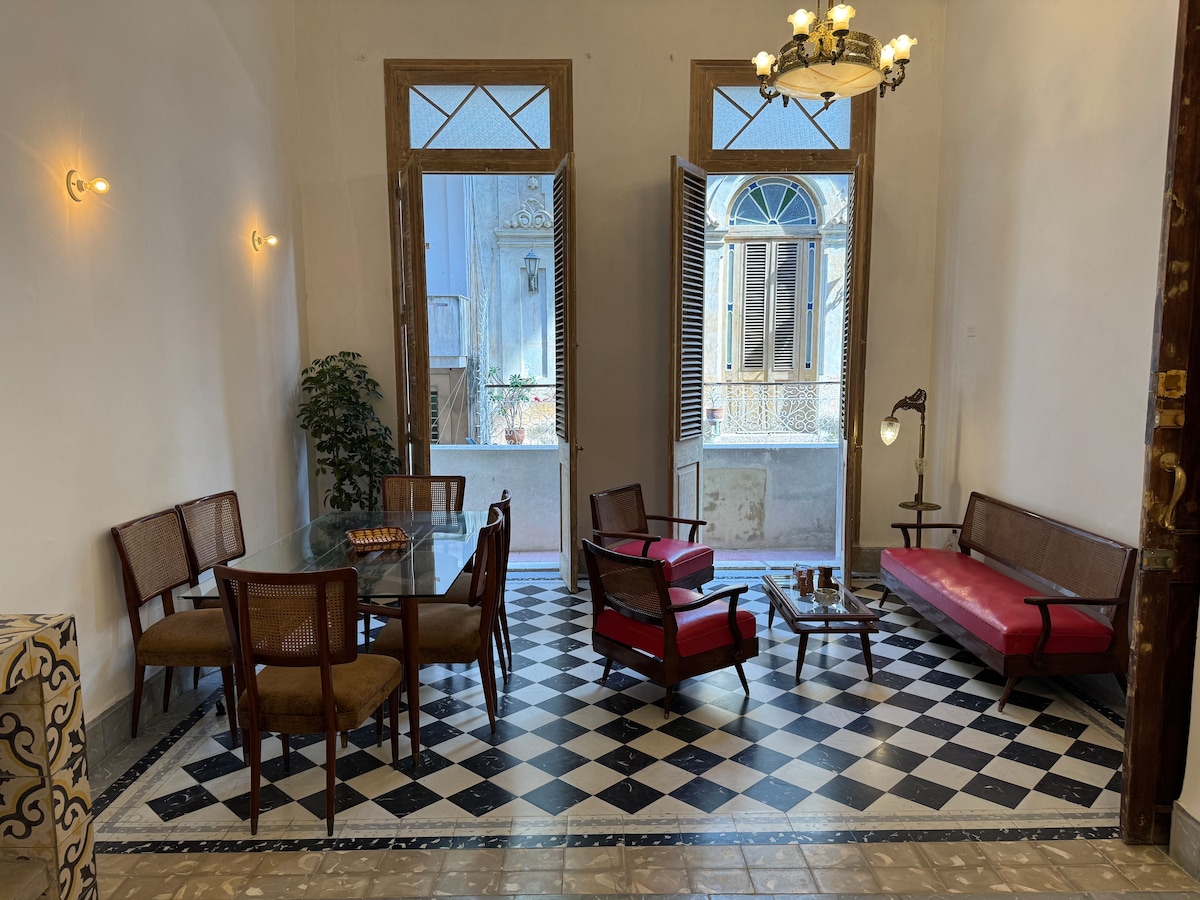 A spacious living area features a combination of modern and vintage furniture. A glass dining table is accompanied by wooden chairs, while a red sofa and armchairs invite relaxation. Light pours in through open doors, enhancing the room's charm with patterned tile flooring.