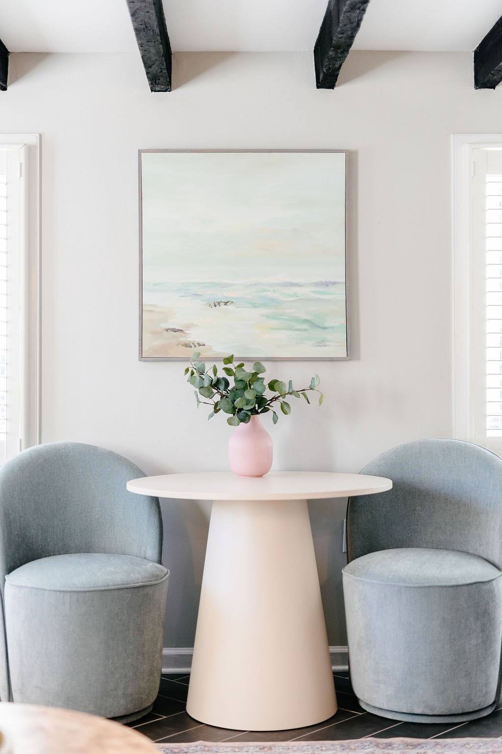 A small dining space features a round table with a sculptural base, paired with two cushioned chairs in soft colors. A pink vase filled with greenery sits at the center, complemented by a calming ocean-themed painting hanging on the wall, illuminated by natural light from the windows.