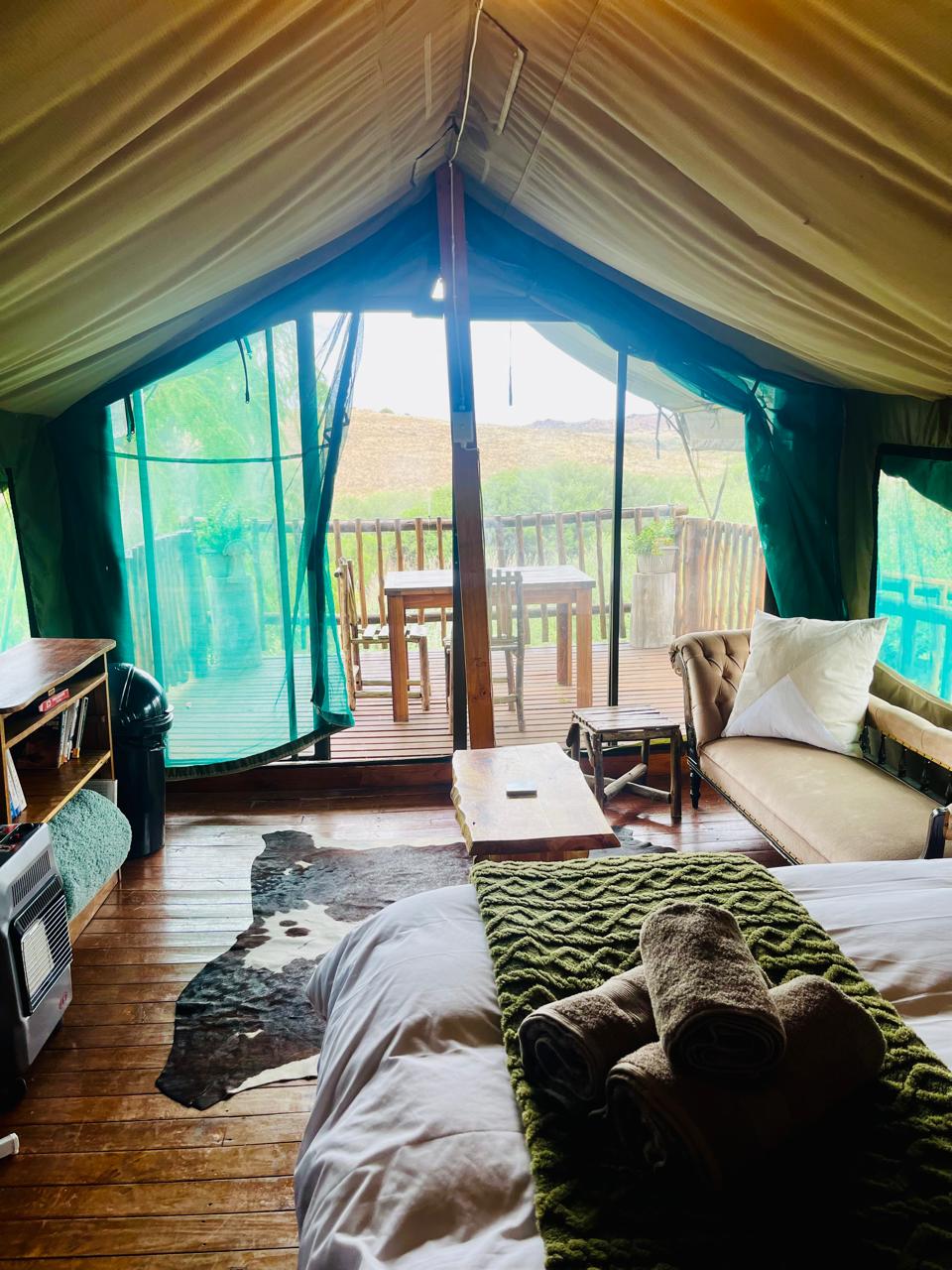 A spacious tent interior features large windows draped in green fabric, allowing natural light to illuminate the area. There is a comfortable bed adorned with a soft blanket and neatly rolled towels. A small wooden table and chairs are visible on the adjacent deck.
