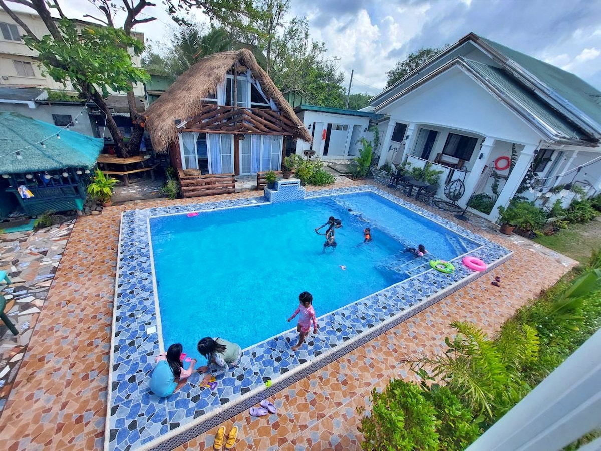 A refreshing pool area features a clear blue pool surrounded by a tiled deck. Children play in the water, enjoying colorful floats. Nearby, two thatched-roof accommodations and a well-maintained garden complete the vibrant outdoor space, offering a relaxed atmosphere for families.