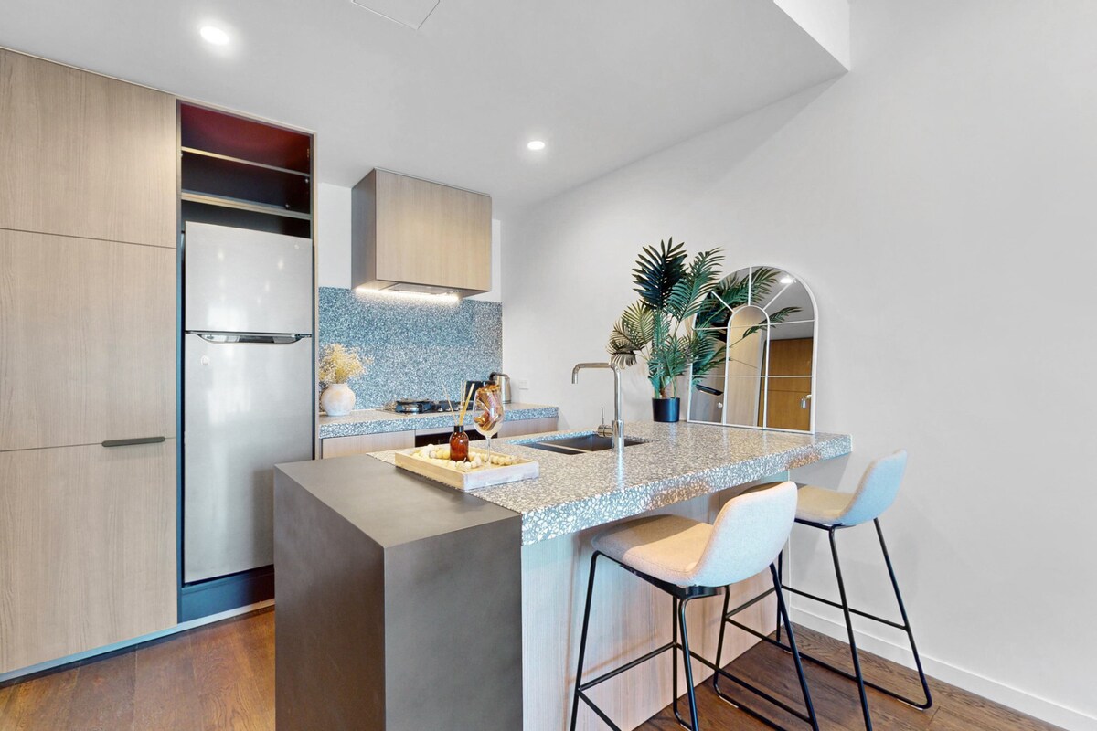 The kitchen features a modern design with light wood cabinetry and a spacious countertop. Two stylish bar stools are positioned at the counter. A large mirror and decorative plants add elements of interest, while appliances are neatly integrated into the cabinetry.