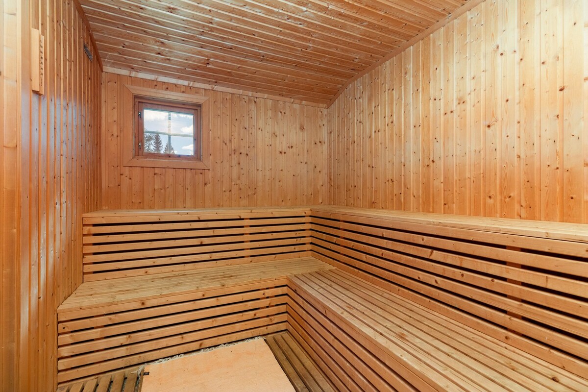 A wooden sauna interior features warm timber paneling and neatly arranged wooden benches. A small window allows natural light to filter in, highlighting the cozy atmosphere while enhancing the rustic design. The floor is covered with a simple mat, completing the tranquil space.