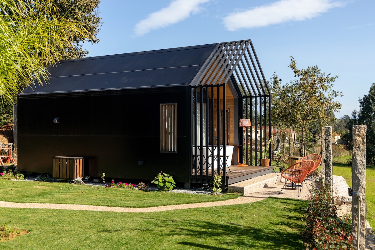 A triangular-shaped villa features a dark exterior contrasted by large windows and a welcoming porch. Surrounding greenery is complemented by flower beds. Outdoor seating is arranged on the porch, inviting relaxation amidst the tranquil garden setting.