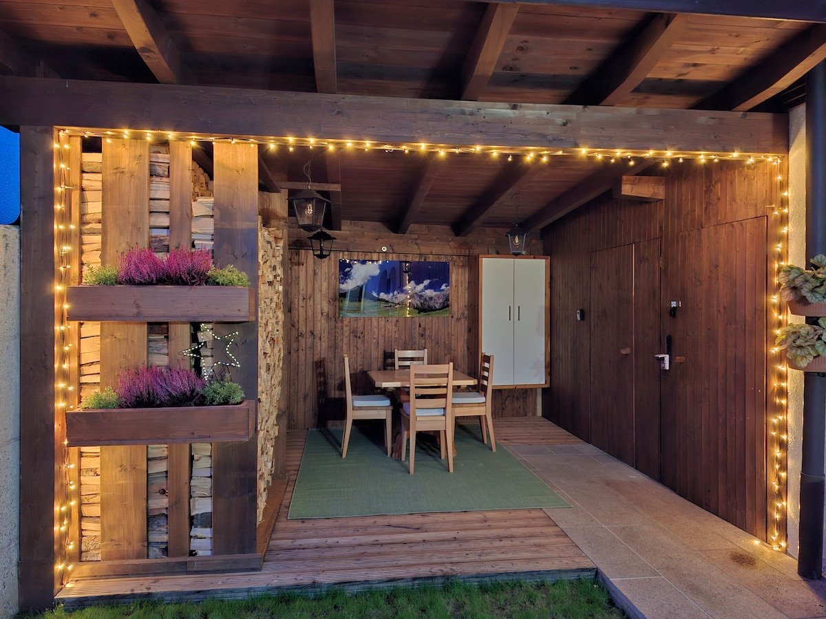 An outdoor terrace area is illuminated by warm string lights, creating an inviting atmosphere. A wooden table with matching chairs is positioned on a green mat, surrounded by planters filled with grass and flowers. Rustic wooden walls enhance the charming ambiance.