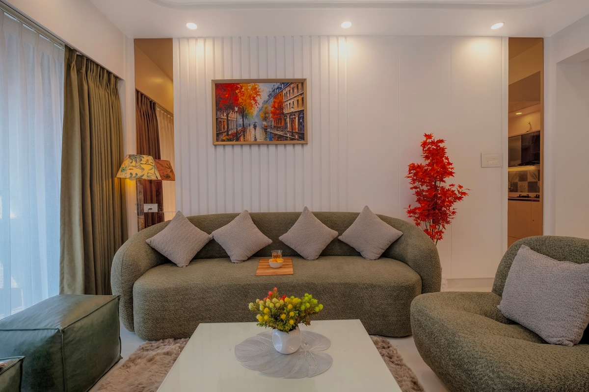 A modern living area is furnished with a curved green sofa, adorned with plush gray cushions. A glass coffee table sits center stage, complemented by a floral arrangement. A vibrant artwork depicting a cityscape hangs above the sofa, and a decorative tree adds a touch of warmth.