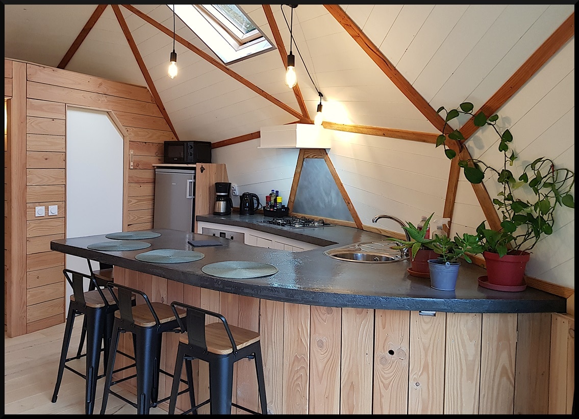 The kitchen area features a curved wooden bar with four metal stools, providing a social space for meals. Natural light filters through a skylight, highlighting the modern appliances and ample cabinetry. Potted plants add greenery to the inviting atmosphere.