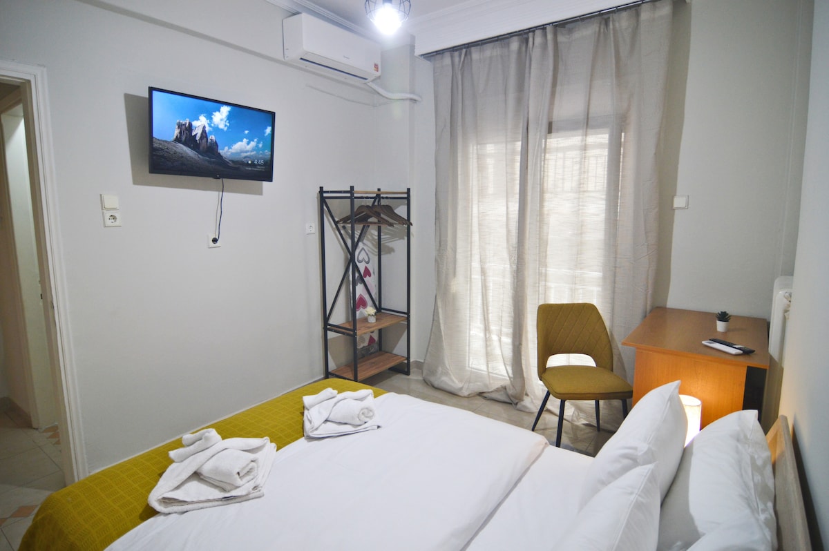 A well-organized sleeping area is displayed featuring a spacious bed with neatly arranged linens. A television is mounted on the wall opposite the bed. A window is dressed with light curtains, allowing natural light to brighten the room. A small desk and chair provide a functional workspace.