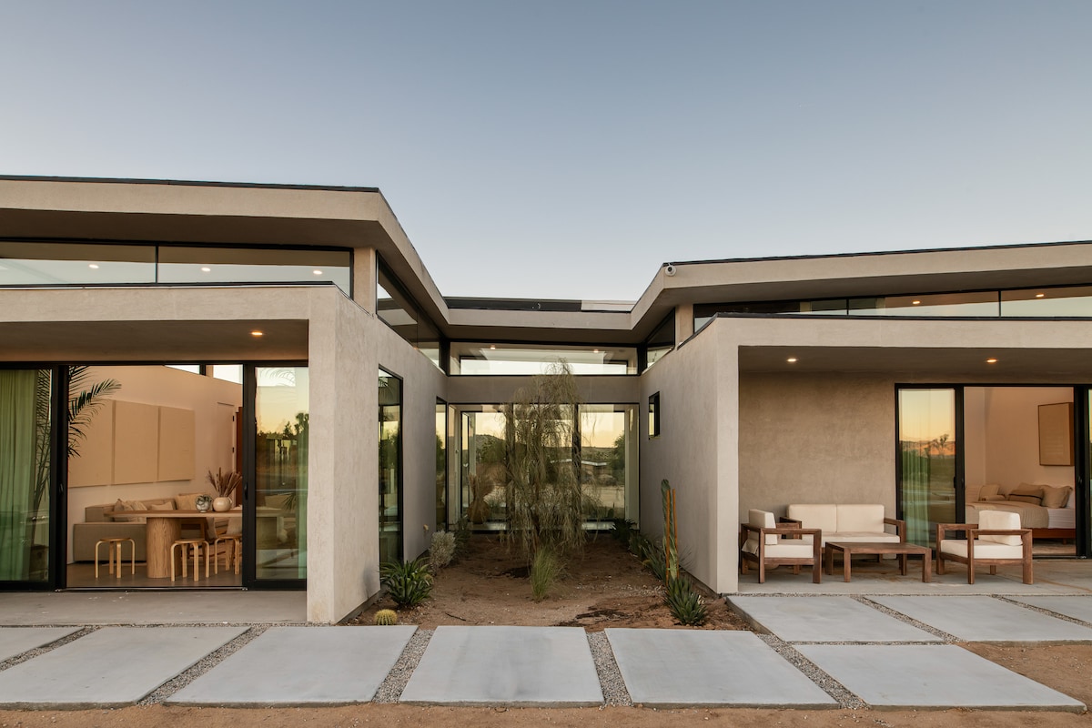 A modern desert home is showcased with clean lines and expansive glass windows. The structure features an open courtyard area with minimal landscaping, highlighting the natural desert environment. The interior spaces are visible through the glass, suggesting a seamless connection with the outdoors.