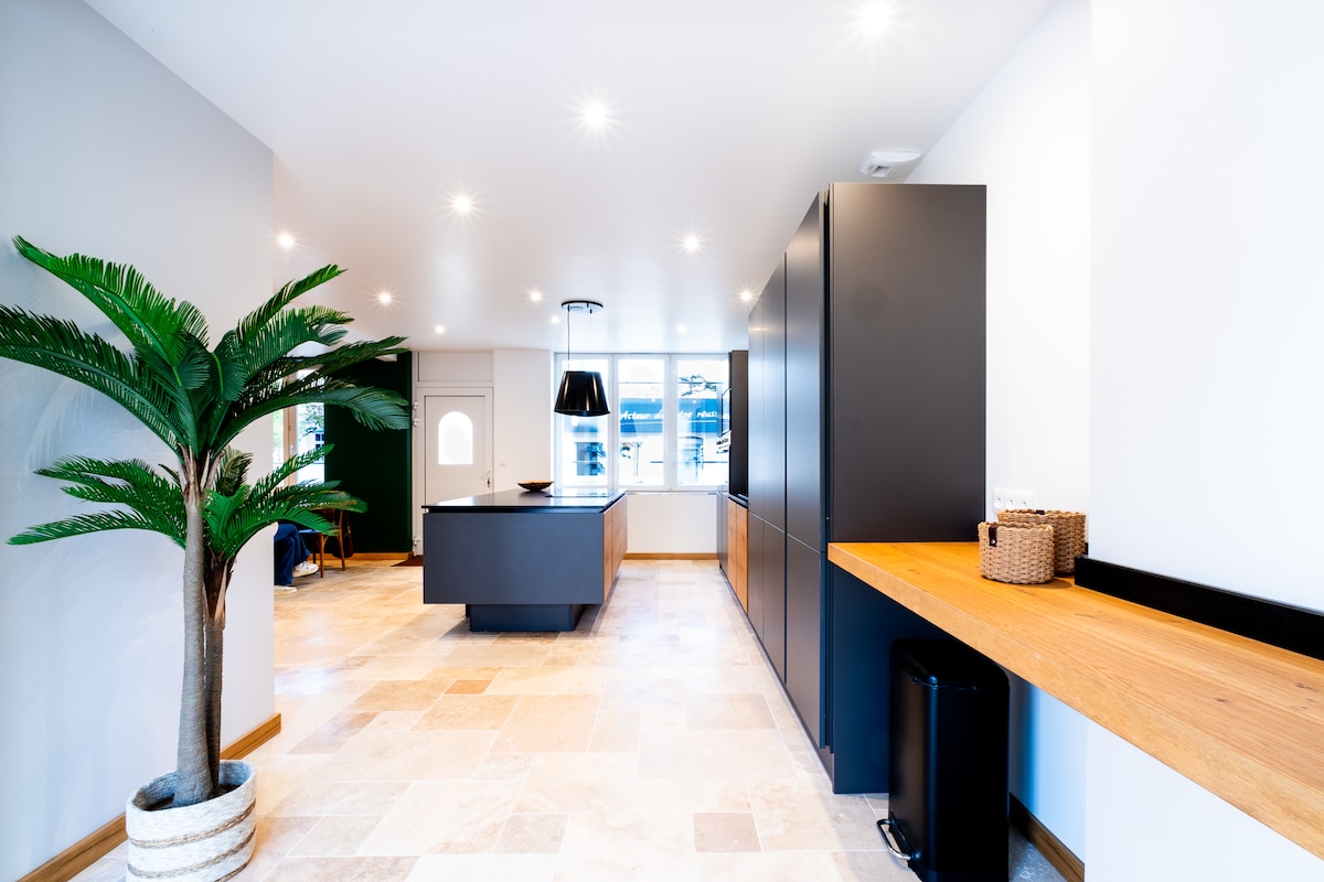 The kitchen space features sleek, modern cabinetry in a muted tone, complemented by a large island at the center. Natural light streams through the window, illuminating the light-colored stone floor and a potted plant. A minimalistic pendant light hangs above the island, enhancing the clean design.