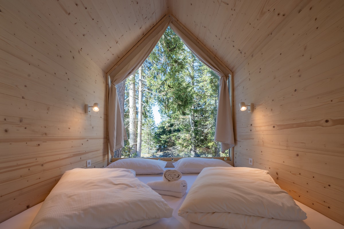 A spacious sleeping area features a large double bed with soft pillows, set against a backdrop of natural wood walls. Large windows framed by light curtains provide views of the surrounding trees, allowing ample natural light to fill the space.