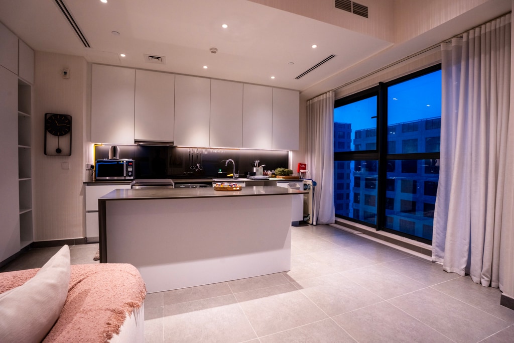 A modern kitchen space is illuminated with soft lighting. Equipped with sleek cabinetry and a central island, it features essential appliances, including a microwave and oven. Large windows provide a view of the surrounding area while sheer curtains allow for natural light.