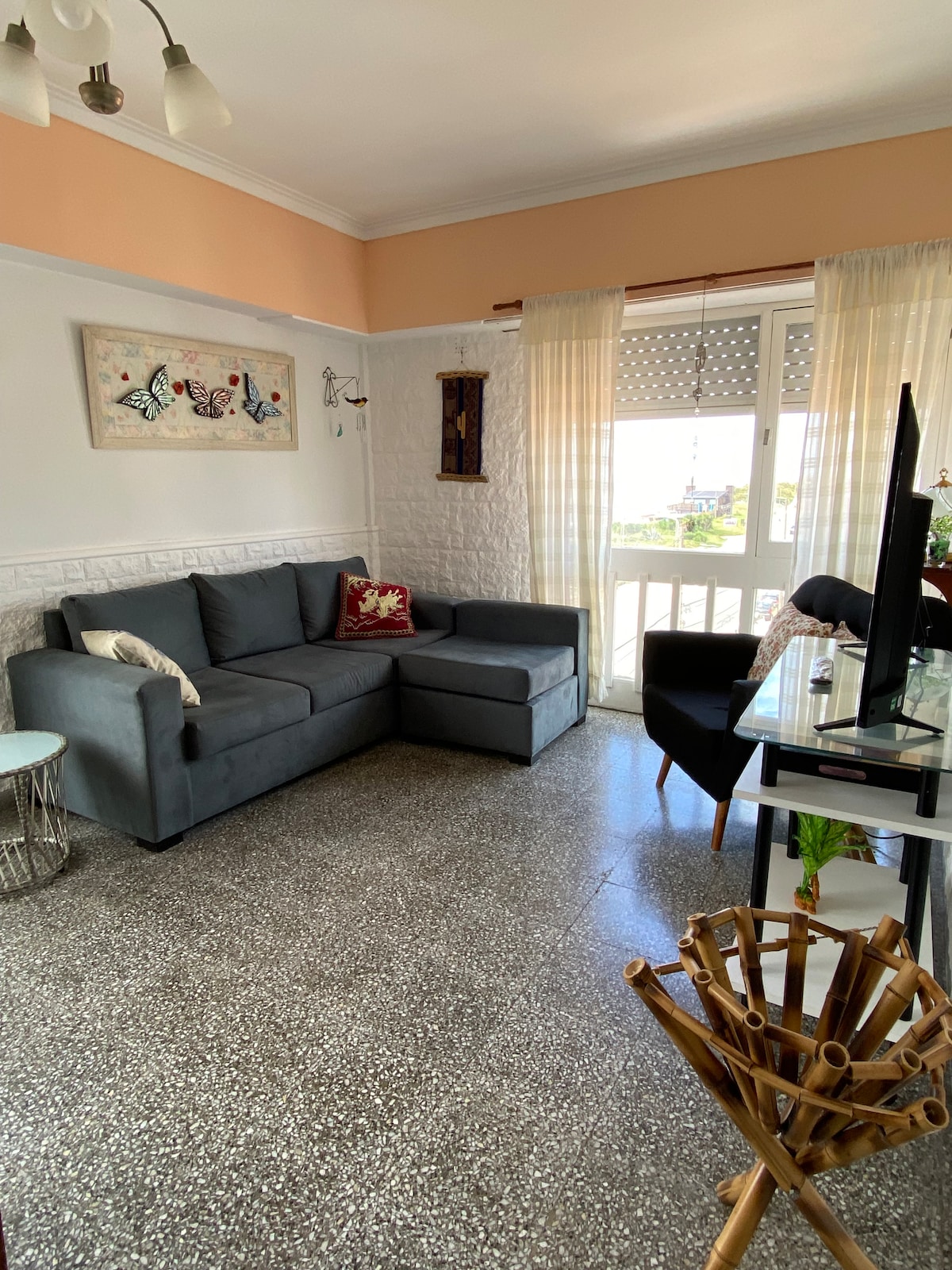 A living area features a gray sectional sofa and a glass-top coffee table. A black desk with a chair is positioned near a window, allowing natural light to illuminate the space. Decorative artwork is displayed on the wall, adding subtle character to the room.
